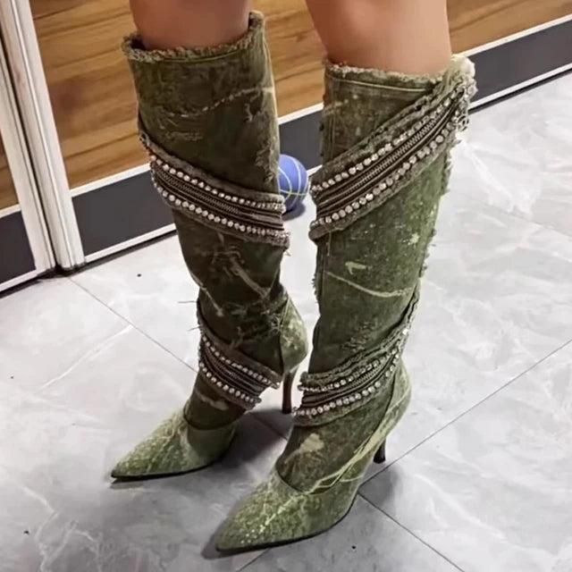 Olive green denim knee-high boots featuring a distressed style, stiletto heel, pointed toe, and zippered accents with rhinestones.