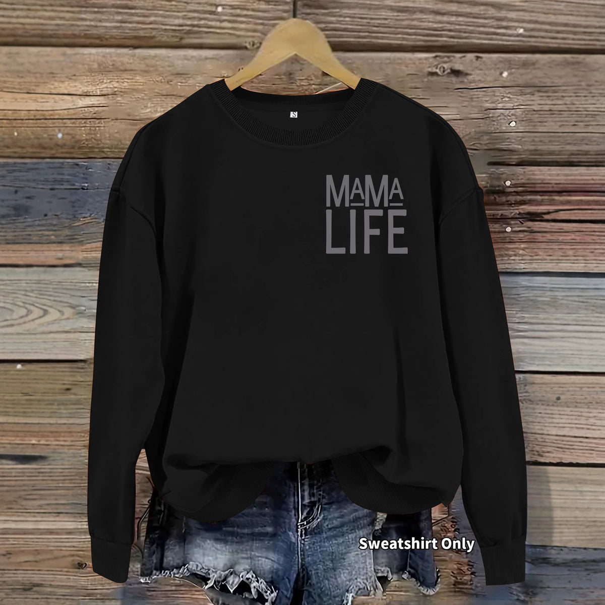 Black crewneck sweatshirt with "MAMA LIFE" text on the chest, hanging on a wooden background.