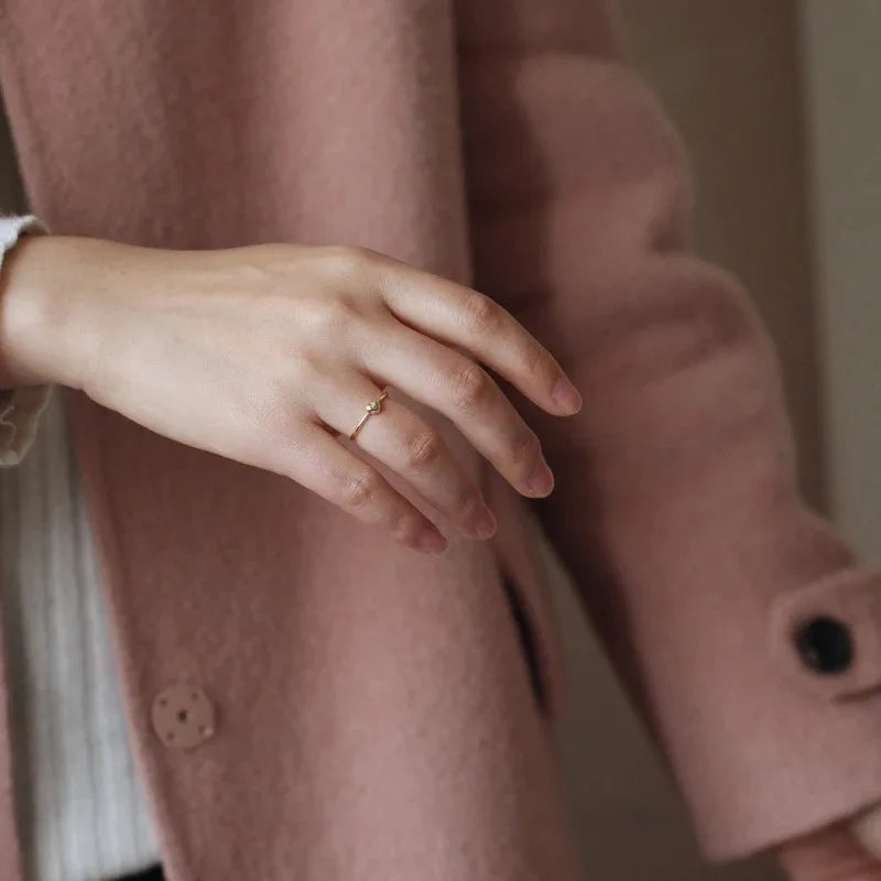 A thin gold ring with a small knot detail on a woman's finger, against a pink wool coat.