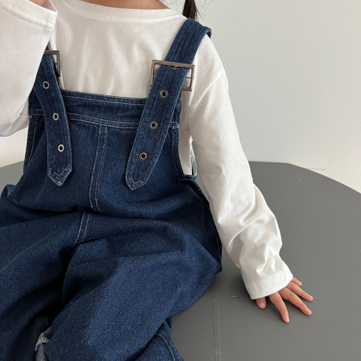 Dark blue denim overalls featuring adjustable straps, metal buckles, and white stitching, worn over a white long-sleeve shirt.