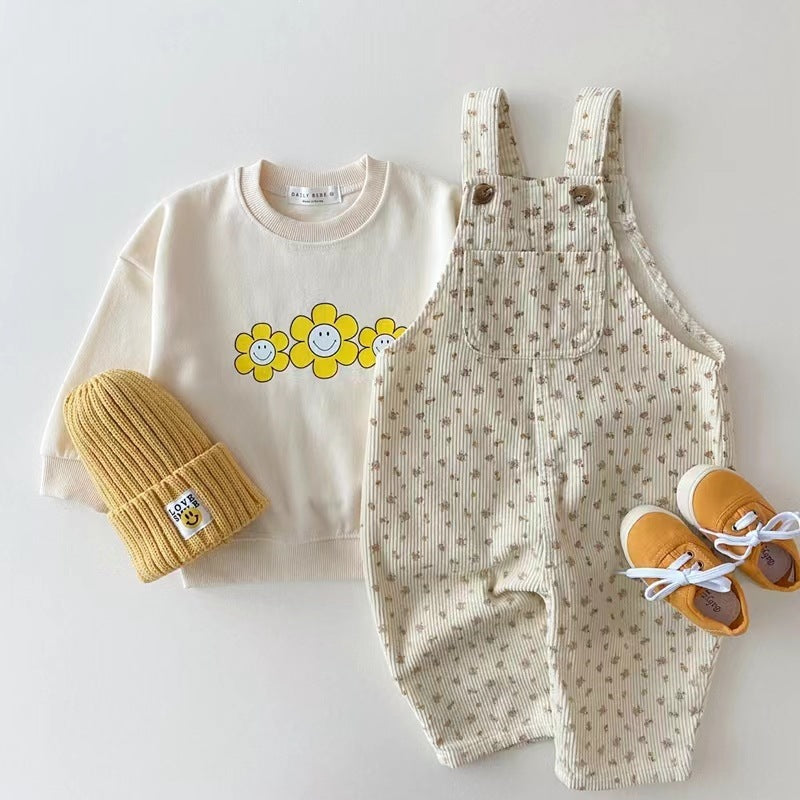 Toddler's outfit: cream sweatshirt with happy flower print, floral corduroy overalls, mustard beanie, and yellow shoes.