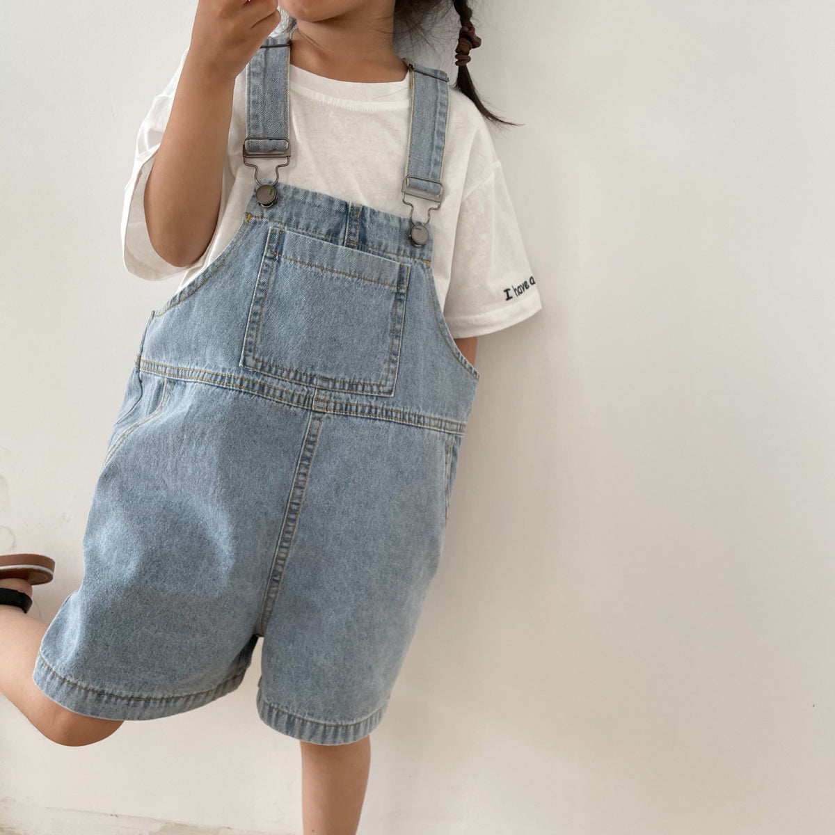 Light blue denim overall shorts with metal clasp straps and a front pocket, worn over a white T-shirt.