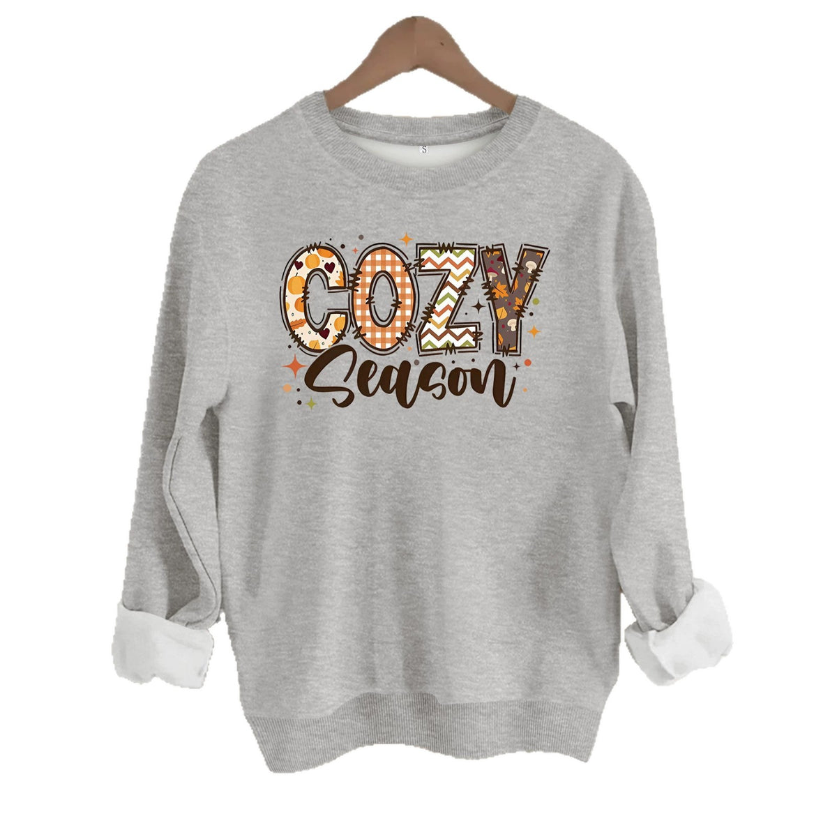 Gray cotton crewneck sweatshirt featuring a patchwork "COZY" and script "Season" graphic in fall colors on a hanger.
