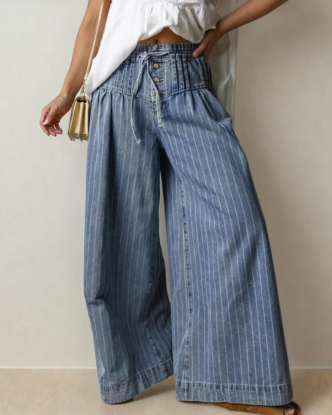 A person models blue striped wide-leg jeans with a high pleated waistband, buttons, and a drawstring.
