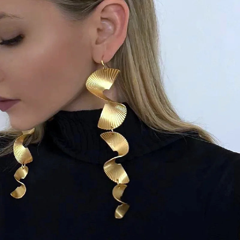 Long gold metal earrings featuring a fluted fan-shaped spiral pattern, worn by a woman.