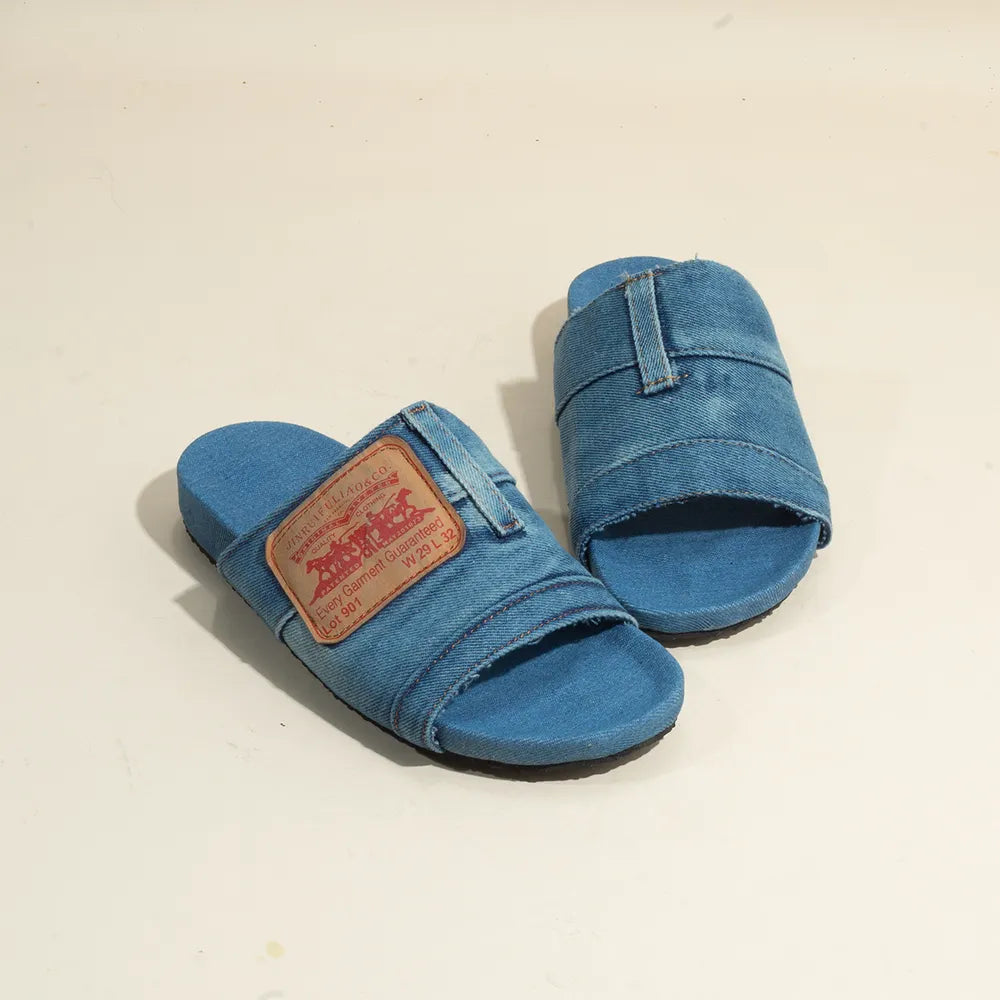 A pair of blue denim slide sandals with a brown leather logo patch on the left strap.
