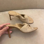 Beige knit stiletto slide sandals with a knotted bow detail and textured heel, held by a hand.