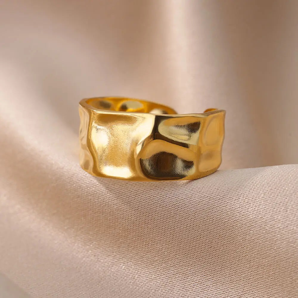 Gold metal ring with organic, melted texture and open back on a soft beige fabric background.