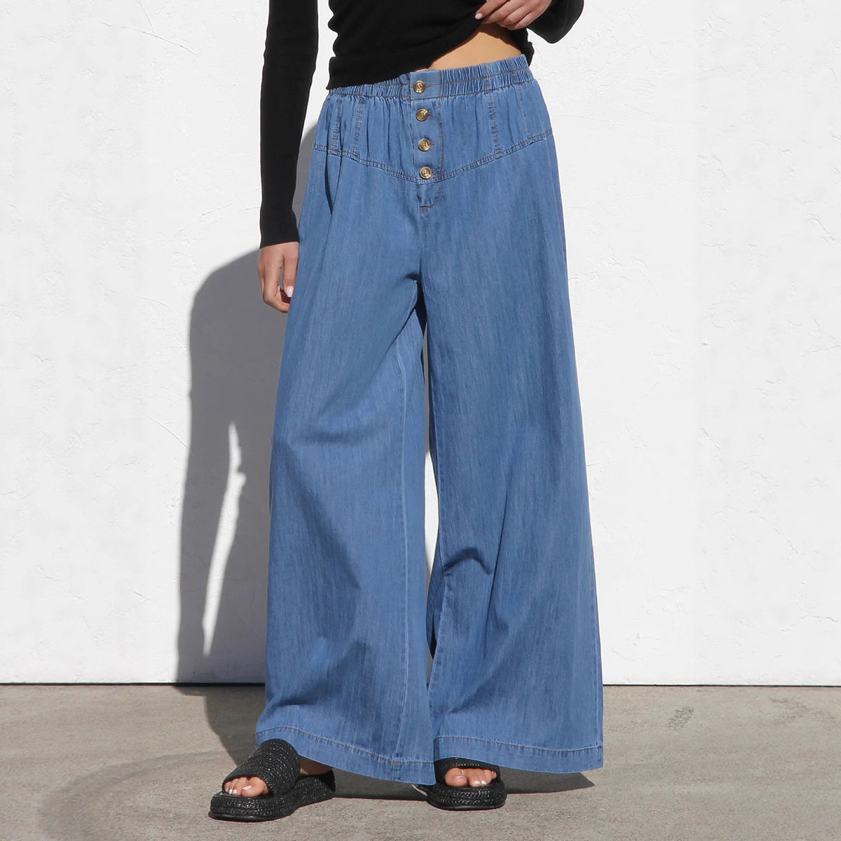 Blue wide-leg denim pants featuring an elastic waist and decorative buttons, paired with a black long-sleeve top.