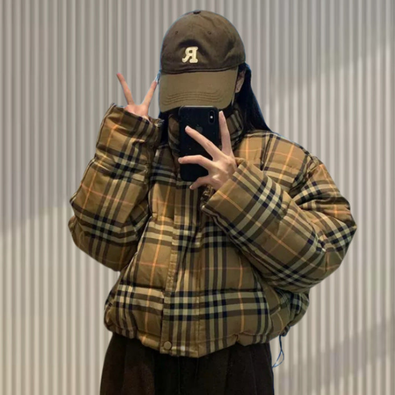A person wears a brown plaid short puffer jacket, a matching brown cap, and dark pants while taking a mirror selfie.