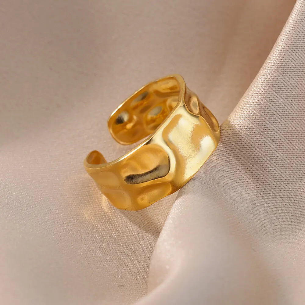 Wide, gold metal ring with an uneven, hammered surface, resting on folded beige fabric.