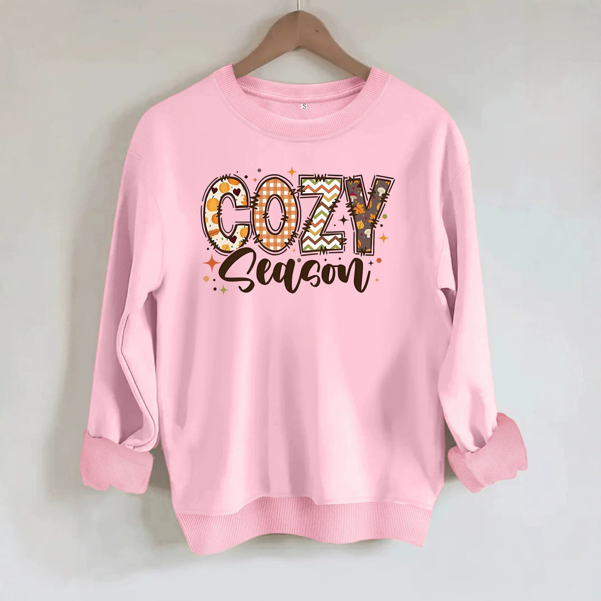 Light pink crewneck sweatshirt with "COZY season" graphic in autumn colors and patterns, hanging on a hanger.