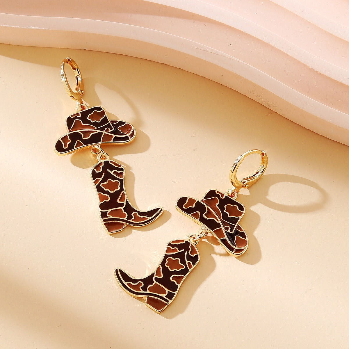 Gold dangle earrings featuring enamel cow print cowboy hats and boots in brown and white.