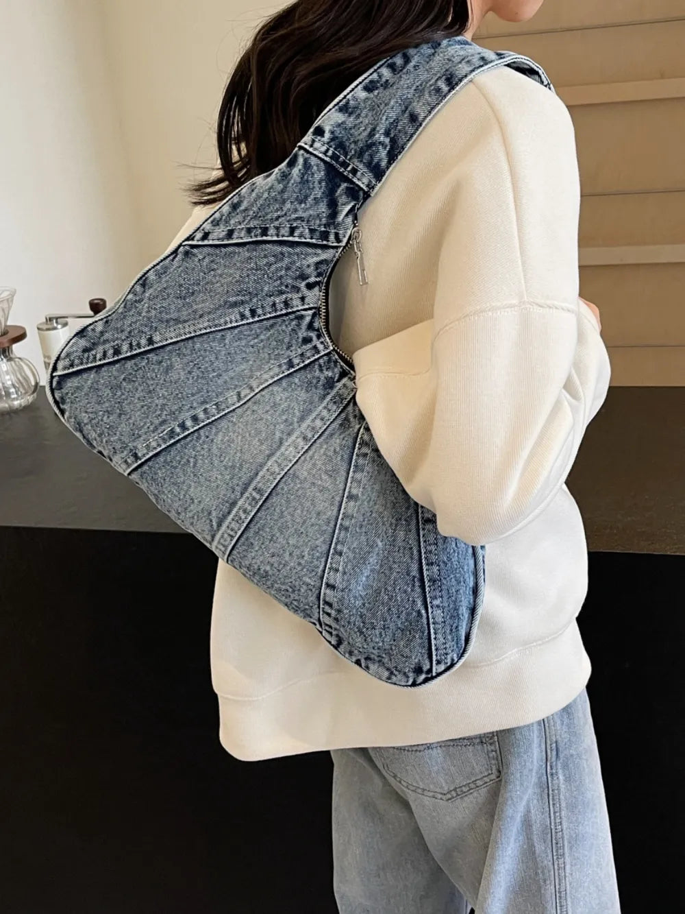 Blue denim croissant-shaped shoulder bag with panel seams and a zipper closure worn by a person.