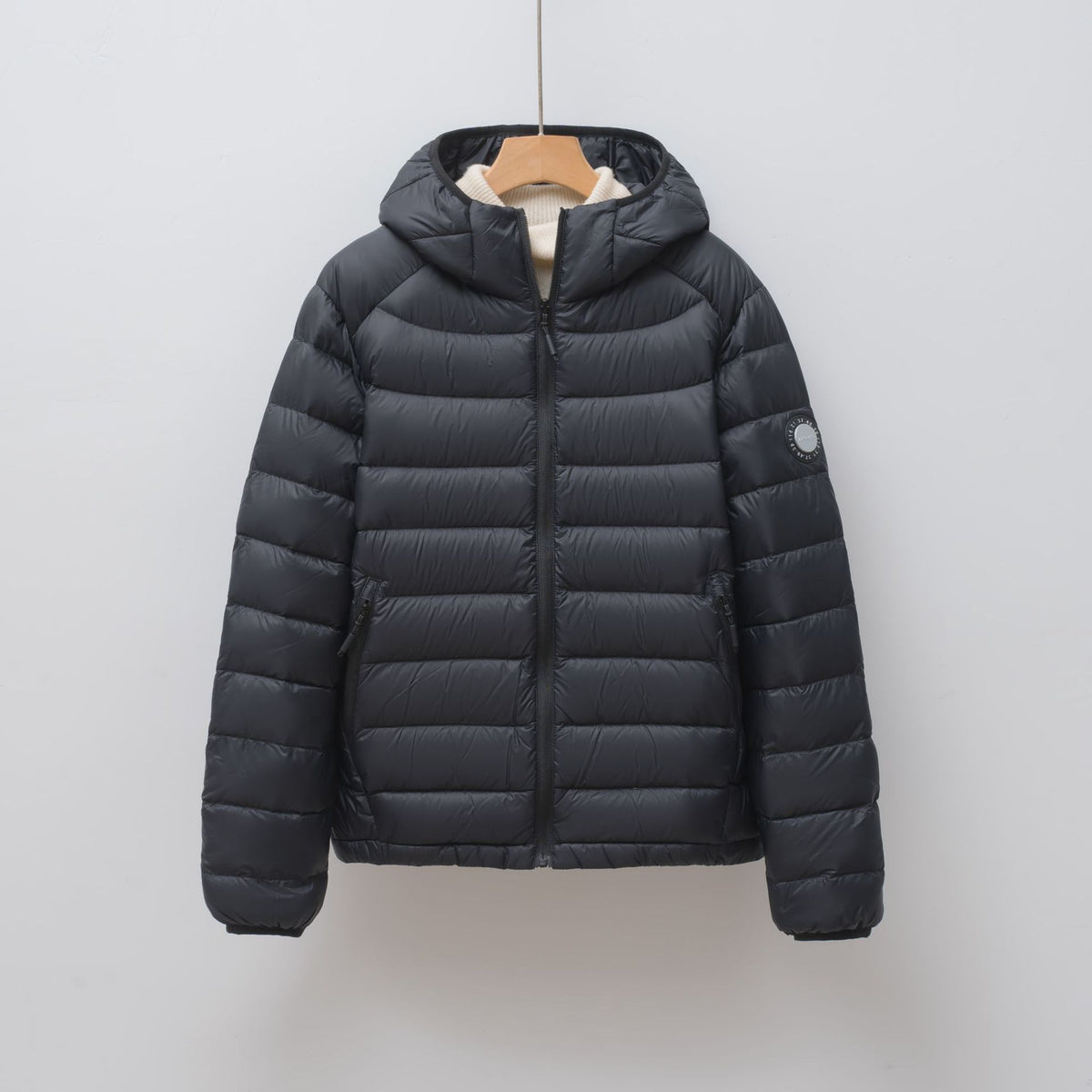 Black puffer jacket with a hood, horizontal quilting, zipper, and a round sleeve patch hanging on a hanger.