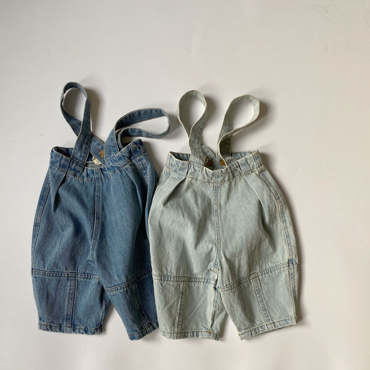 Two pairs of denim overalls in dark and light blue, featuring button straps and pleated front details.