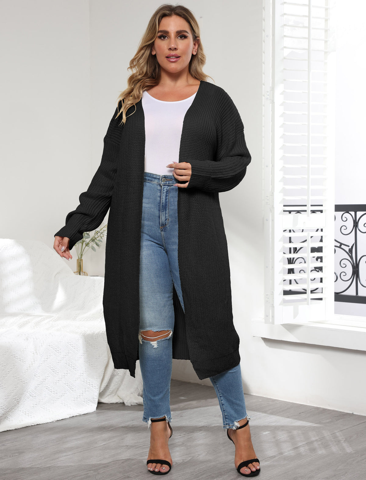 A plus-size model wears a long black knit cardigan over a white top and ripped blue jeans.