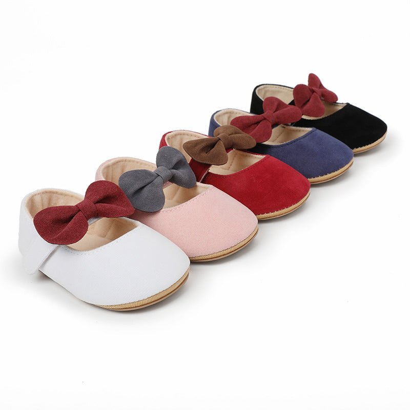 Five solid-color faux suede baby shoes with contrasting bows, in white, pink, red, blue, and black.