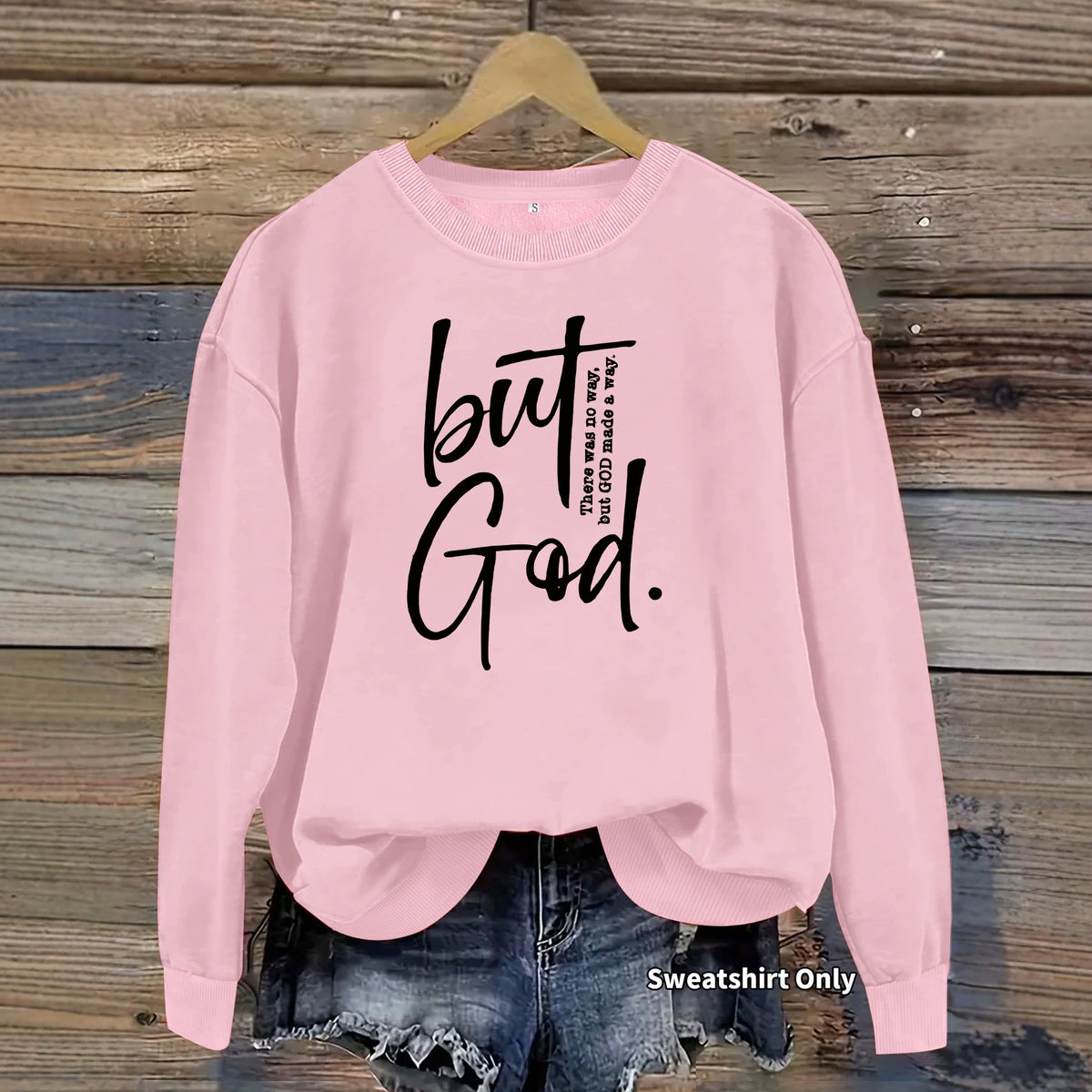Pink cotton crewneck sweatshirt with black "But God" text on the chest, hanging against a wooden background.
