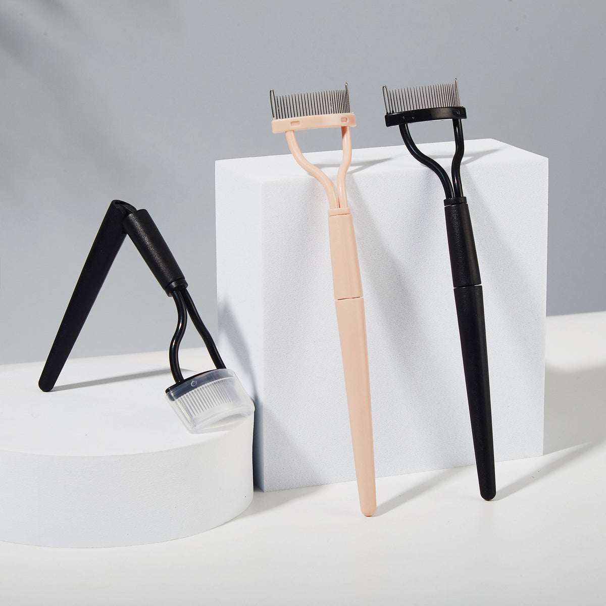 Three plastic eyelash separator tools, one pink and two black, featuring metal teeth and protective caps.