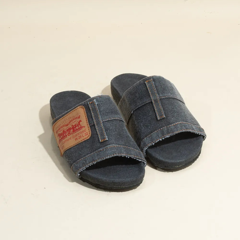 Dark blue denim slide sandals with thick straps, contrast stitching, frayed edges, and a brown leather patch.