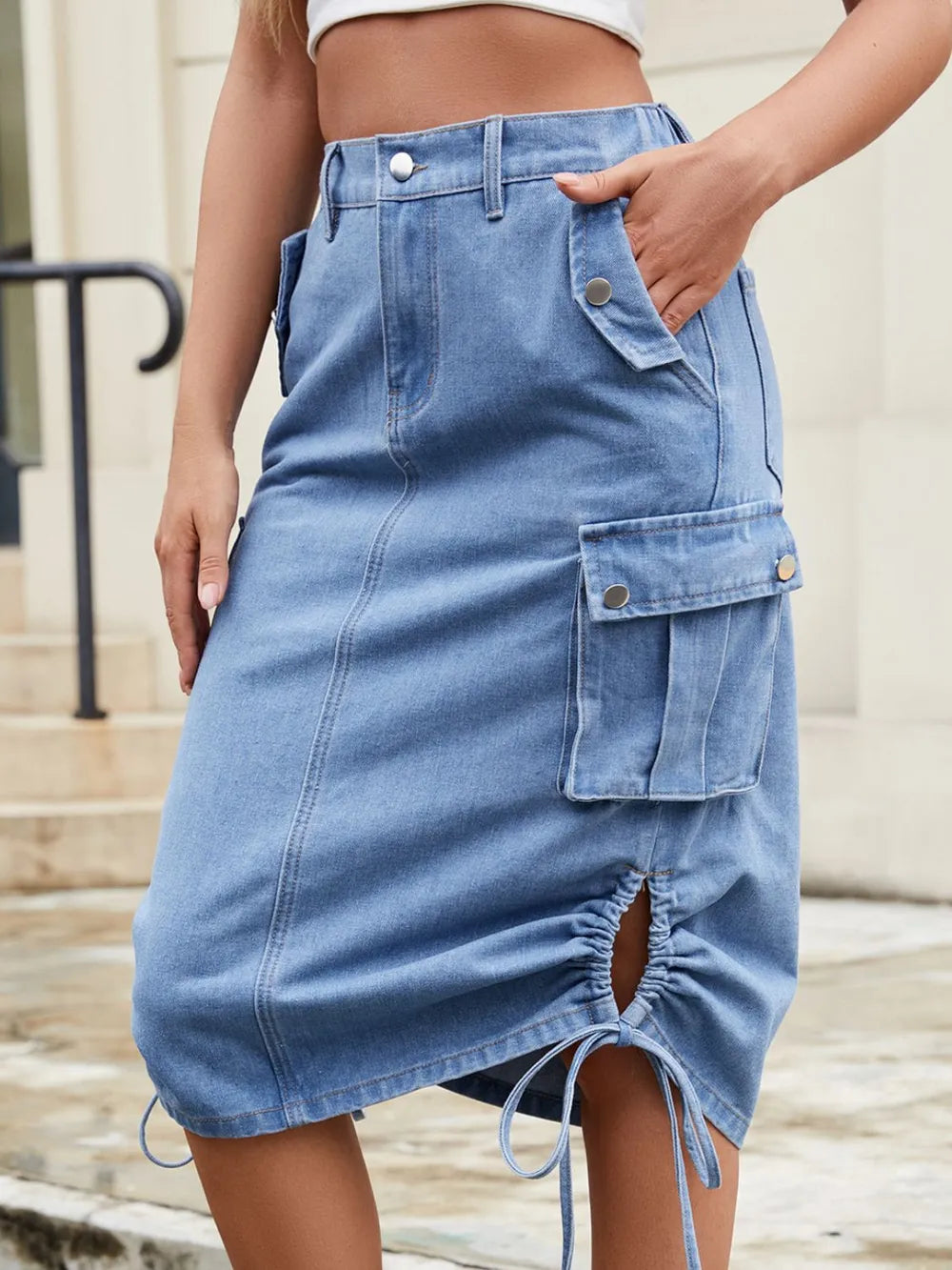 Light blue denim cargo skirt with large flap pockets, side drawstrings, front slit, and button waist.