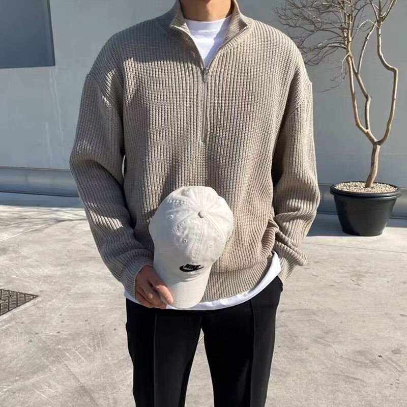 Beige ribbed knit quarter-zip sweater with high collar, worn over white t-shirt, holding white cap.