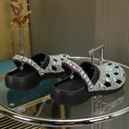 Black platform clogs featuring iridescent rhinestone embellishments and a pivoting heel strap on a glass table.