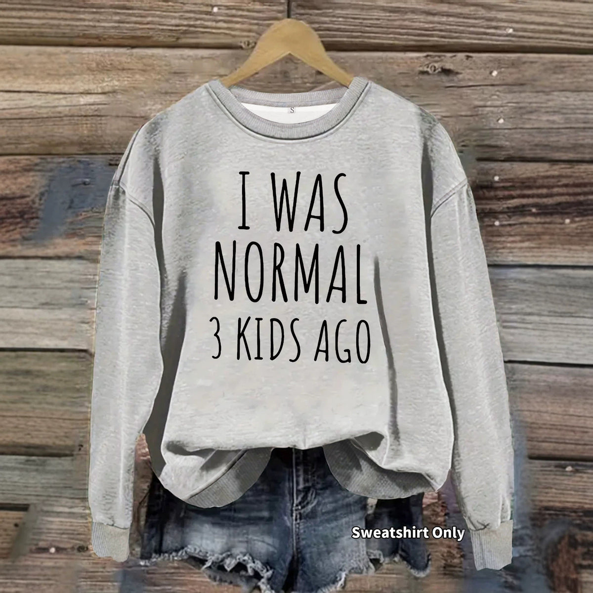 Gray sweatshirt with black text reading "I WAS NORMAL 3 KIDS AGO" hanging on a wooden wall.