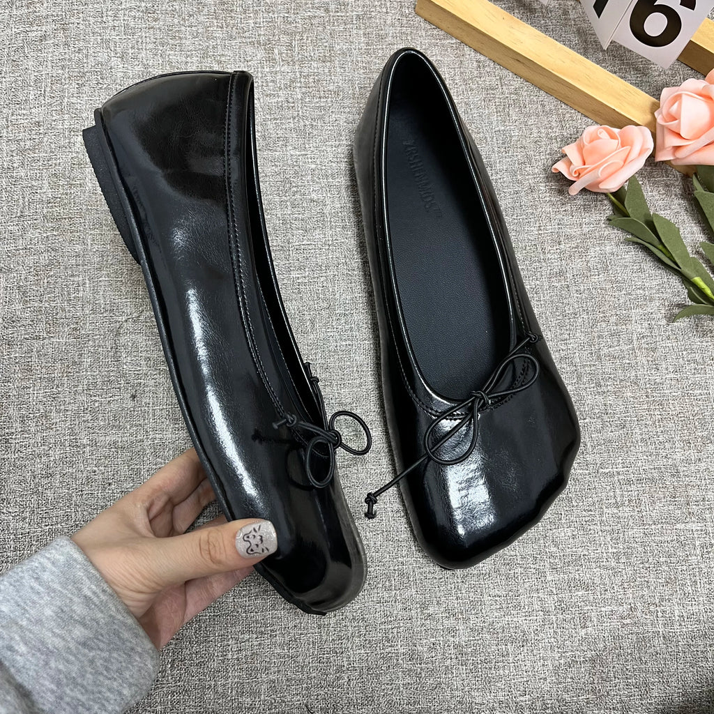 Sophisticated black glossy slip-on flats with a round toe and delicate bow detail, held by a hand against a gray fabric.