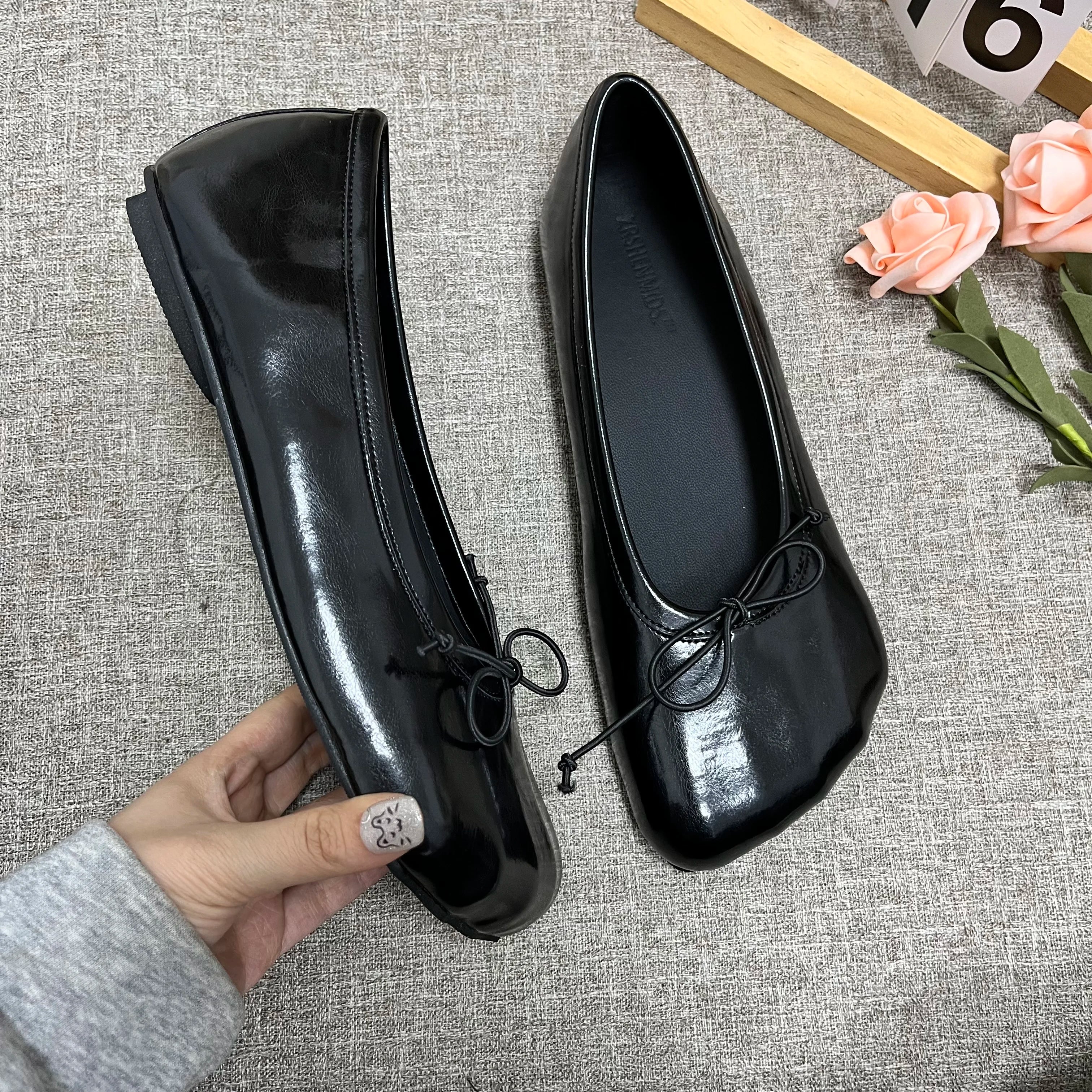 Sophisticated black glossy slip-on flats with a round toe and delicate bow detail, held by a hand against a gray fabric.