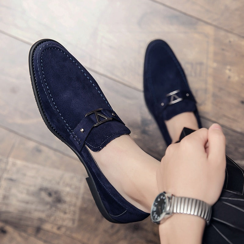 Dark blue suede slip-on loafers with a metal V-strap buckle, topstitching, and a wooden floor background.