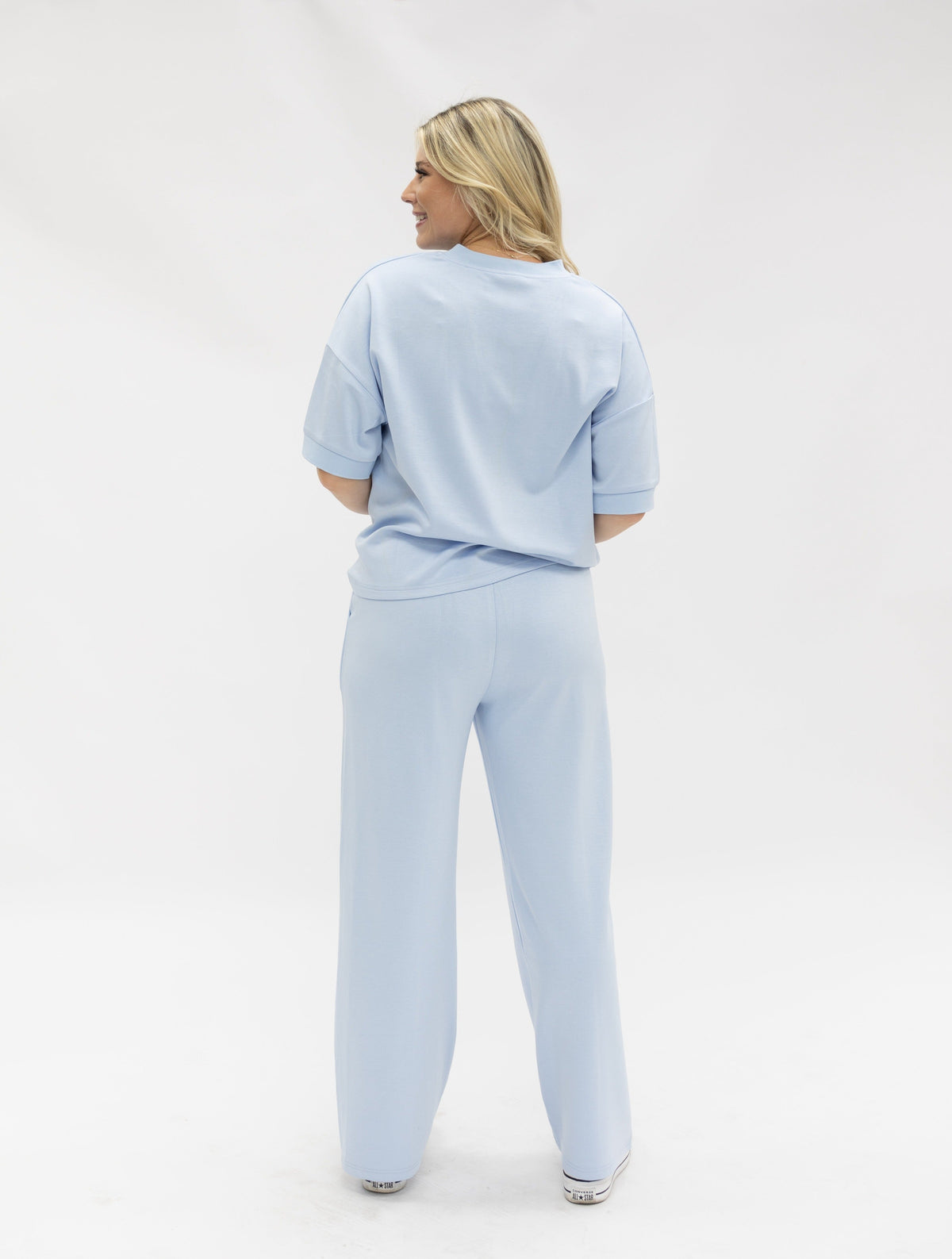 A blonde woman stands facing away, wearing a light blue short-sleeved top and matching long pants.