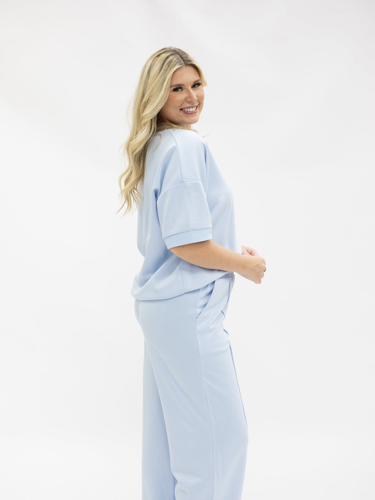 A blonde woman stands against a white background, wearing matching light blue pants and a short-sleeved top.
