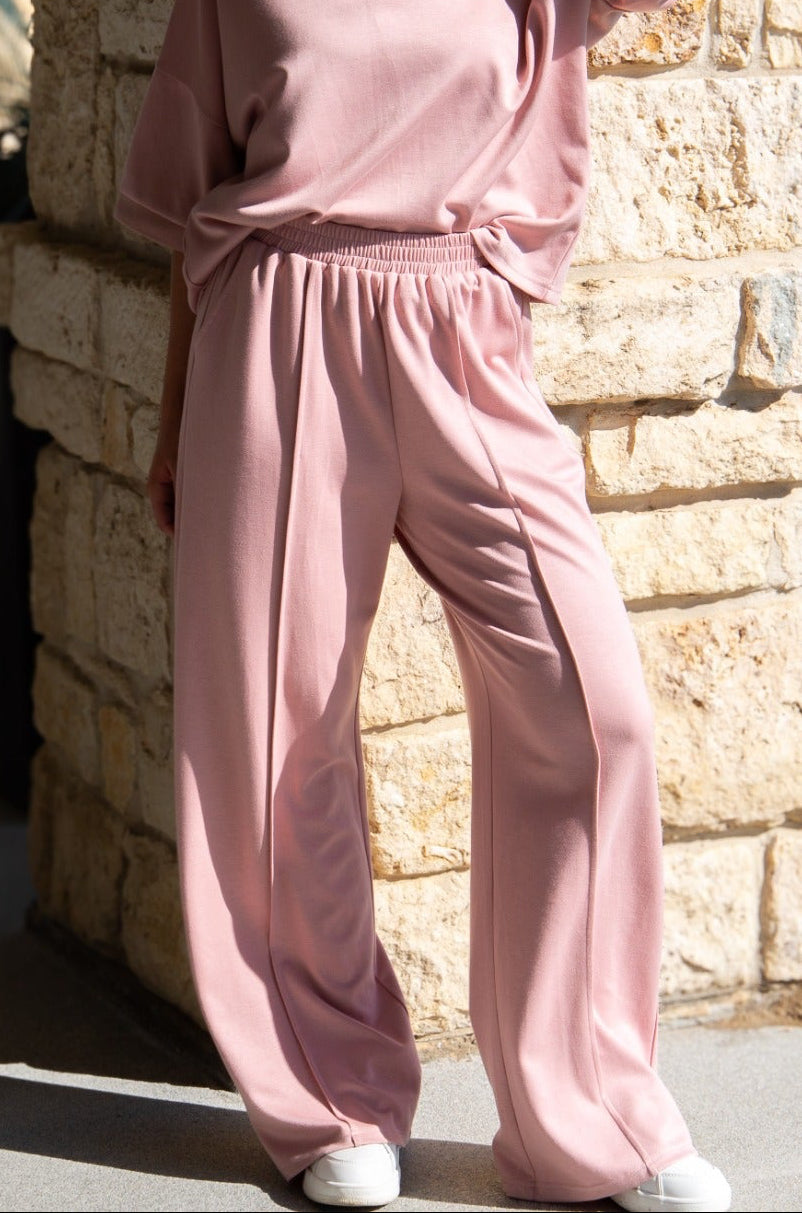 A person wearing wide-leg pink pants with center seams and a matching top against a stone wall.