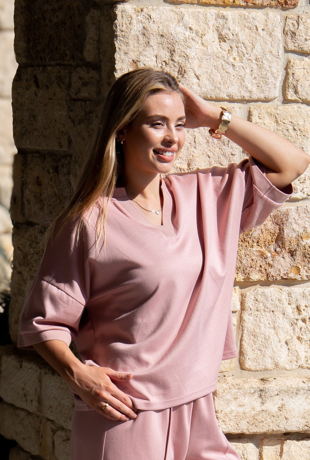 A woman poses in a solid pink V-neck top with short sleeves, leaning against a stone wall.